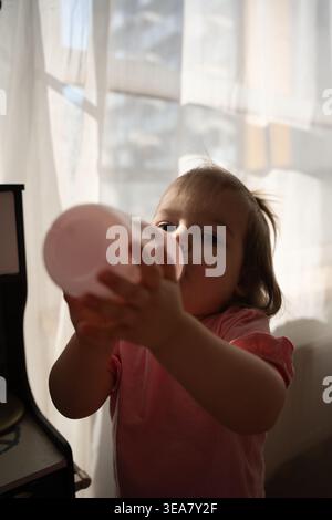 Junge Kleinkinder im warmen Morgenlicht trinken aus einer rosafarbenen Flasche an transparenten Vorhängen in einer gemütlichen Wohnung, offenes Nahporträt mit weichen Schatten, Gen Stockfoto