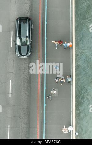 Ein Hochwinkelblick fängt den täglichen Rhythmus des städtischen Lebens ein, während die Menschen auf einem Bürgersteig neben Stadtverkehr und Wasser spazieren. Stockfoto