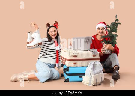 Nettes junges Paar mit Laptop, Koffern und kleinem Weihnachtsbaum auf beigefarbenem Hintergrund. Winterurlaubskonzept Stockfoto