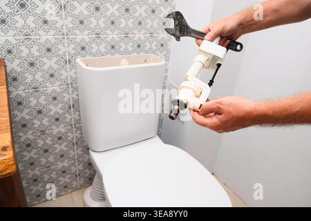 Nahaufnahme der Hände eines Handwerkers, der ein Ersatzventil für die Toilettenbefüllung und einen Schraubenschlüssel hält, während er an der Zisterne arbeitet. Hauswartung und Sanitär-Reparatur Stockfoto