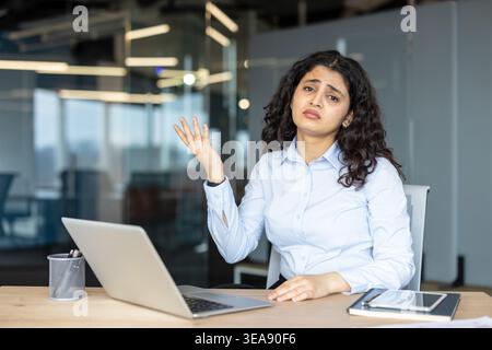 Die junge indische Frau fühlt sich frustriert und unsicher, zuckt mit einer gleichgültigen Geste, während sie in die Kamera blickt, sitzt mit einem Laptop an ihrem Schreibtisch in einer modernen Büroumgebung Stockfoto