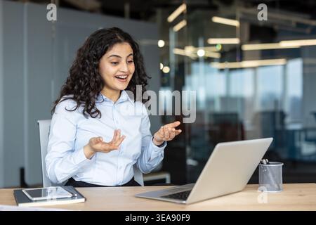 Junge indische Geschäftsfrau, die ein virtuelles Meeting auf einem Laptop in einem modernen Büro hat, während sie mit Gesten an einem Schreibtisch kommuniziert und ihre Arbeit diskutiert Stockfoto