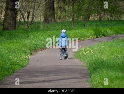 Rückansicht eines kleinen Jungen in einer blauen Jacke, der auf einem gewundenen Asphaltweg in einem Frühlingswald oder Park auf einem Balance Bike fährt. Hellgrünes Gras auf der Straße Stockfoto