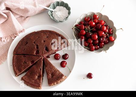 Rustikale Gourmet-Komposition mit einem runden Schokoladenkuchen mit gebackenen Kirschen, in Portionen auf einem weißen Teller geschnitten. Stockfoto