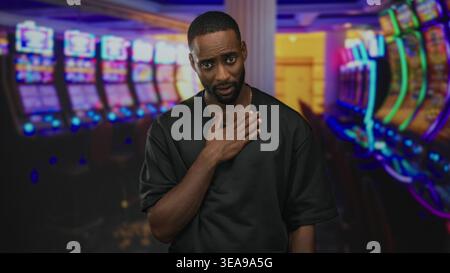 Junger afroamerikanischer Mann mit Hand auf der Brust in der Nähe von beleuchteten Spielautomaten im Eingangsbereich des Casinogebäudes; bedauern Reue-Entschuldigung. Stockfoto