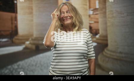 Eine ältere Frau zeigt den Finger auf den Tempel, während sie lächelt, ein gestreiftes Hemd trägt und an Steinsäulen im Gebäude steht; Zufriedenheit reflektiert. Stockfoto