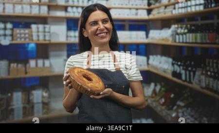Bäckerin hält einen runden Kuchen mit Händen, lächelt und lacht, während sie eine Schürze in einem Ladengebäude trägt; Freude Gastfreundschaft Wärme. Stockfoto