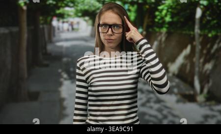 Frau mit Brille und gestreiftem Hemd zeigt mit dem Finger auf den Tempel auf der sonnendurchfluteten Straße mit schattigen Schatten; nachdenkliche Konzentration. Stockfoto