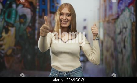 Frau, die in einer Graffiti-Straßenallee Daumen mit sichtbaren Daumen hochlegt und ein breites Lächeln trägt, in Creme-Pullover und Jeans, mit Blick auf die Kamera; Glück Stockfoto