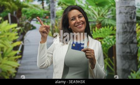 Frau, die die Kreditkarte hält, zeigt mit dem Finger nach oben auf der Straße mit tropischen Pflanzen und einer Promenade dahinter; Selbstvertrauen stärken. Stockfoto