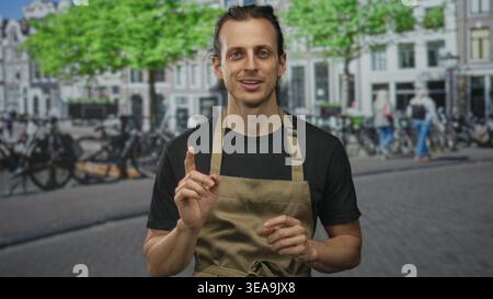 Junger Mann mit Händen, die Palmen auf der Straße hochgehoben haben, mit Schürze und schwarzem Hemd, langen Haaren, die zurückgebunden sind; Entschuldigung Demut. Stockfoto