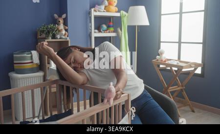 Die Frau legt den Kopf auf das Gittergeländer, übergibt die Augen, hält rosafarbenes Spielzeug im Babyzimmer mit Wickeltisch und Fenster; Pflegemüdigkeit. Stockfoto