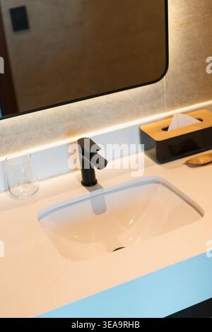 Modernes weißes Waschbecken befindet sich unter einem großen Spiegel. Schwarzer Wasserhahn sorgt für einen eleganten Touch. Klares Glas liegt neben dem Waschbecken. In der Nähe befindet sich ein hölzerner Taschentuch. LED li Stockfoto