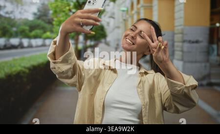 Hispanische Frau in einem lässigen Hemd, das Smartphone auf Armlänge hält, ein Friedensschild macht und lächelt auf einer Stadtstraße in der Nähe eines Steingebäudes; verspieltes c Stockfoto