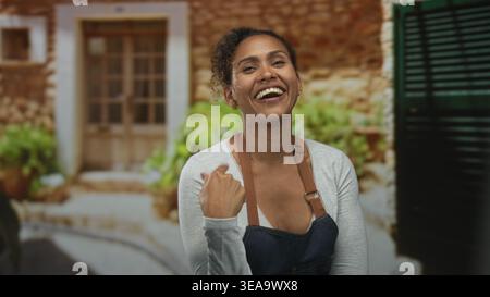 Frau, die mit dem Finger auf die Kamera im Gebäudeeingang zeigt, Schürze trägt und mit gekreuzten Armen lächelt; warmes Selbstvertrauen. Stockfoto