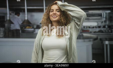 Frau, die Haare mit der Hand berührt, leichtes Lächeln und geschlossene Augen in einer Restaurantküche; ruhige Besinnung. Stockfoto