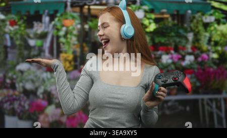 Rothaarige Frau mit Gamepad und Katzenohrkopfhörern, Handbewegung auf einem Straßenblumenmarkt; verspielte Freude. Stockfoto