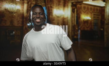 Ein Mann lächelt, trägt ein weißes T-Shirt mit Ohrring und sichtbaren Dreadlocks, steht in einem reich verzierten Museumsgebäude mit warmer Beleuchtung; freudiges Vertrauen Stockfoto
