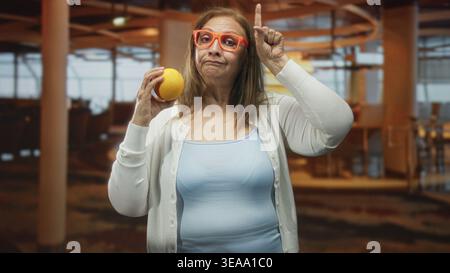 Frau, die Orange in der Hand hält und mit dem Finger auf den Tempel im Flughafenterminal zeigt, mit orangefarbener Brille und hellblauem Tanktop; bewundernswerte Neugier. Stockfoto