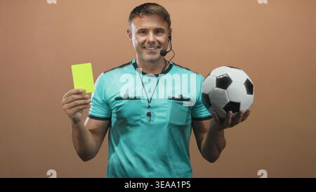 Mann Schiedsrichter hält gelbe Karte in der Hand und Fußball, trägt türkisfarbenes Schiedsrichtertrikot, Headset und Pfeife im Studio; Vertrauen Fairness. Stockfoto