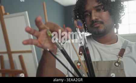 Junger indianer hält Pinsel zwischen den Fingern und inspiziert Borsten im Studio, während er subtil lächelt; kreative Konzentration. Stockfoto