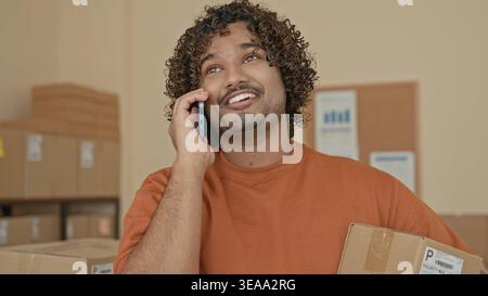 Mann mit Pappkarton und Smartphone, Hand an Ohr beim Lächeln und Reden, umgeben von Paketen im Gebäude; Optimismus für kleine Unternehmen. Stockfoto