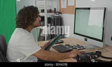 Man Fotograf tippt im Studio neben Kamera und Desktop-Computer an einem Holztisch mit Monitor und Tastatur auf ein Tablet; fokussierte Kreativität. Stockfoto