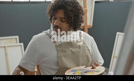 Mann, junger inder, mit beigefarbener Schürze, mit hölzerner Farbpalette und Pinsel, während er im Kunststudio Gesten auf Leinwand streckt; fokussierte Kreativität. Stockfoto