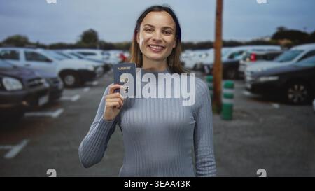 Frau lächelt und hält Pass auf einer Straße mit geparkten Autos auf einem Parkplatz in der Nähe einer Stange und nassem Asphalt; Reisesolz. Stockfoto