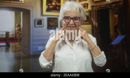Ältere Frau mit Brille zeigt Zeigefinger auf die Wangen, während sie in einem Museumsgebäude zwischen gerahmten Gemälden lächelt; Freude Nostalgie. Stockfoto