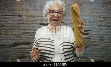 Ältere Frau, die ein langes Baguette in der rechten Hand hält, während sie mit der linken Hand in einem Studio eine Geste zur Kamera drückt; Nostalgie. Stockfoto