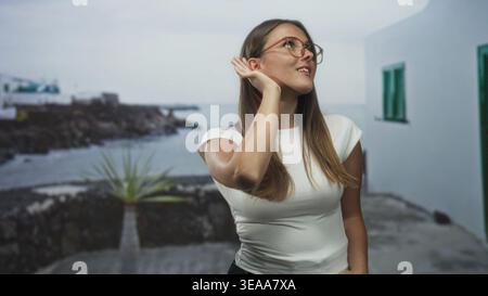 Frau, die ihr Ohr mit der Hand an einem weißen Gebäude am Meer schröpft, Brille und ein weißes Oberteil trägt; neugierige Aufmerksamkeit. Stockfoto