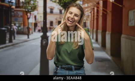 Frau zeigt Zeigefinger auf lächelnde Zähne auf einer sonnigen Straße mit Efeu und Schaufensterfronten; fröhlicher ehrlicher Moment. Stockfoto
