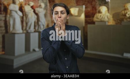 Mann, der den Mund mit Händen in einer Museumsgalerie zwischen klassischen Skulpturen und Steinpodest bedeckt, mit dunklem Hemd und Ringen; Überraschungsreflexion. Stockfoto