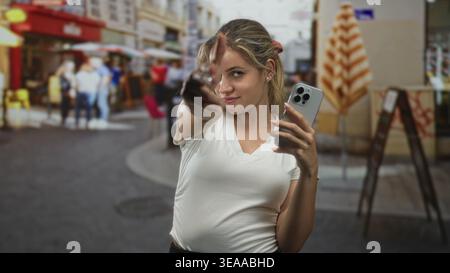 Frau zeigt mit dem Finger auf die Kamera auf der Straße, während sie ihr Smartphone hält und ein weißes T-Shirt trägt; Selbstvertrauen. Stockfoto