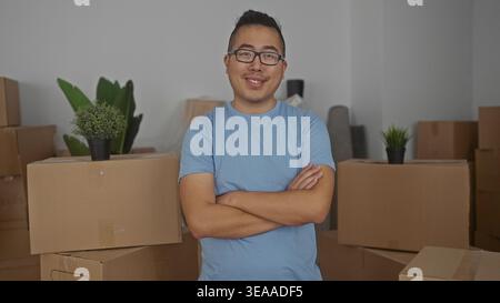 Junger chinesischer Mann mit Armen, die neben gestapelten Pappschachteln im Gebäude während des Umzugs gekreuzt wurden; Zufriedenheit des beweglichen Tages. Stockfoto