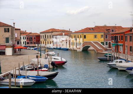 Blick auf den Hauptkanal auf der Insel Murano mit farbenfrohen Gebäuden mit Terrakotta-Dächern, Booten, die an Holzpfählen vertäut sind, einer Ziegelbogenbrücke und Stockfoto