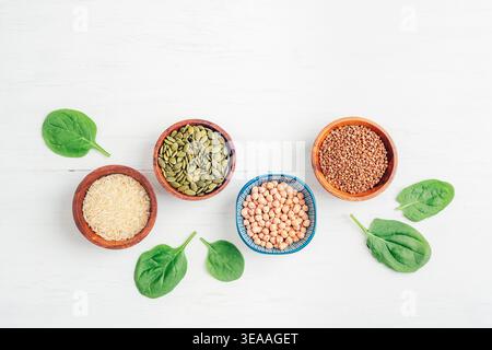 Getreide und Samen in Schüsseln und grüne Spinatblätter auf weißem Holztisch. Reis, Buchweizen, Kichererbsen und Kürbiskerne, gesunde vegane Lebensmittel. Draufsicht, Stockfoto