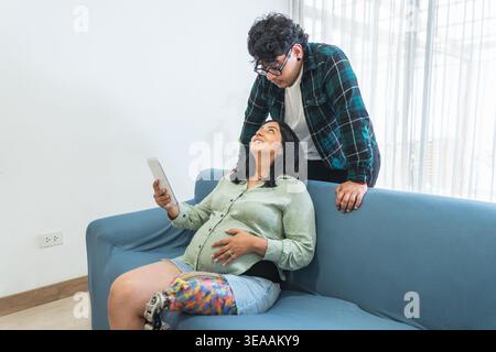 Glückliche schwangere Frau mit einer Beinprothese mit einer Tablette, während ein Mann neben ihr steht Stockfoto