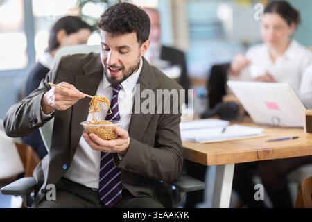 Junger Geschäftsmann, der in der Mittagspause am Arbeitsplatz am Laptop mit anderen Kollegen im Büro zum Mitnehmen essen kann Stockfoto