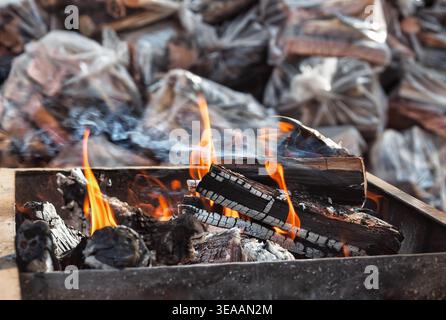 Flammen und weißer Rauch erheben sich aus verkohlten Holzstämmen in einer dunklen Metallfeuergrube mit mehreren Kunststoffsäcken aus Spaltholz im Hintergrund. Stockfoto