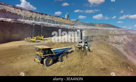 Ein großer gelber Transportwagen wird von einem Bagger in einer tiefen, terrassierten Tagebau unter blauem Himmel mit Erde beladen. Stockfoto