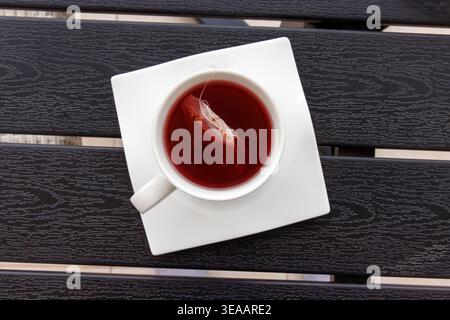 Blick von oben auf eine Tasse roten Früchtetee mit einem Teebeutel in einer weißen Tasse auf einer quadratischen weißen Untertasse, die auf einem dunklen Lattentisch steht. Kopierbereich. Selektiv f Stockfoto