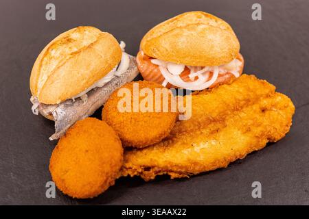 Zwei kleine Sandwiches stehen neben frittierten Fischstücken und Kroketten auf einem dunklen Teller. Die Umgebung scheint während der Nachmittagsstunde ein Lebensmittelmarkt zu sein Stockfoto