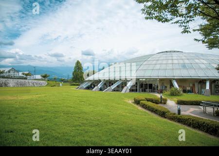 Eine große Glaskuppel befindet sich auf einem breiten grünen Rasen vor einer malerischen Bergkuppel in Japan. Modernes architektonisches Wahrzeichen mit Sommerhimmel und üppiger Umgebung. Stockfoto