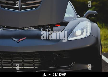 Frontdetail einer mattschwarzen Chevrolet Corvette C7 mit offener Motorhaube, eckigem LED-Scheinwerfer, gekreuztem Flaggenlogo, Frontgrill, aggressiv. Stockfoto