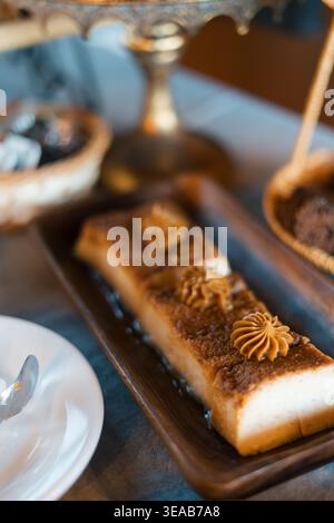 Rechteckiges Dessert ruht auf einem dunklen Holztablett. Es besteht aus mehrschichtigem Kuchen mit cremefarbenen Rosetten. Die weiße Platte hält einen Löffel daneben. Eine weitere Schokolade Stockfoto