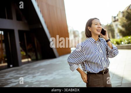 Eine Koreanerin steht vor einem modernen Gebäude. Sie hält ein Handy an ihrem Ohr und lächelt während des Gesprächs. Die Sonne scheint hell auf sie und den Vater Stockfoto