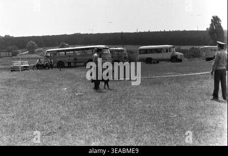 Slawjansk, 1970er Jahre Blick auf ein Hundeübungsfeld während eines Wettkampfes, mit einem Hundeführer, der neben seinem aktiv bellenden Deutschen Schäferhund steht. Der Hintergrund ist gefüllt mit mehreren geparkten Personenbussen aus der Sowjetzeit und Zuschauern, die sich in der Nähe der Baumgrenze versammelten. Dieses atmosphärische Archivbild, das auf authentischem Schwarz-weiß-Film aufbewahrt wird, bietet einen breiteren Blick auf die lokale Sportinfrastruktur und die große Beteiligung an militärisch-technischen Hundeveranstaltungen im friedlichen Donbass. Stockfoto