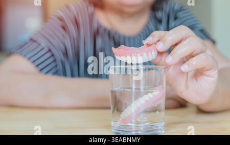 Asiatische ältere oder ältere alte Patientin, die ihre Teilprothese der Ersatzzähne über einem Glas Wasser hält. Nahaufnahme, selektiver Fokus. Stockfoto